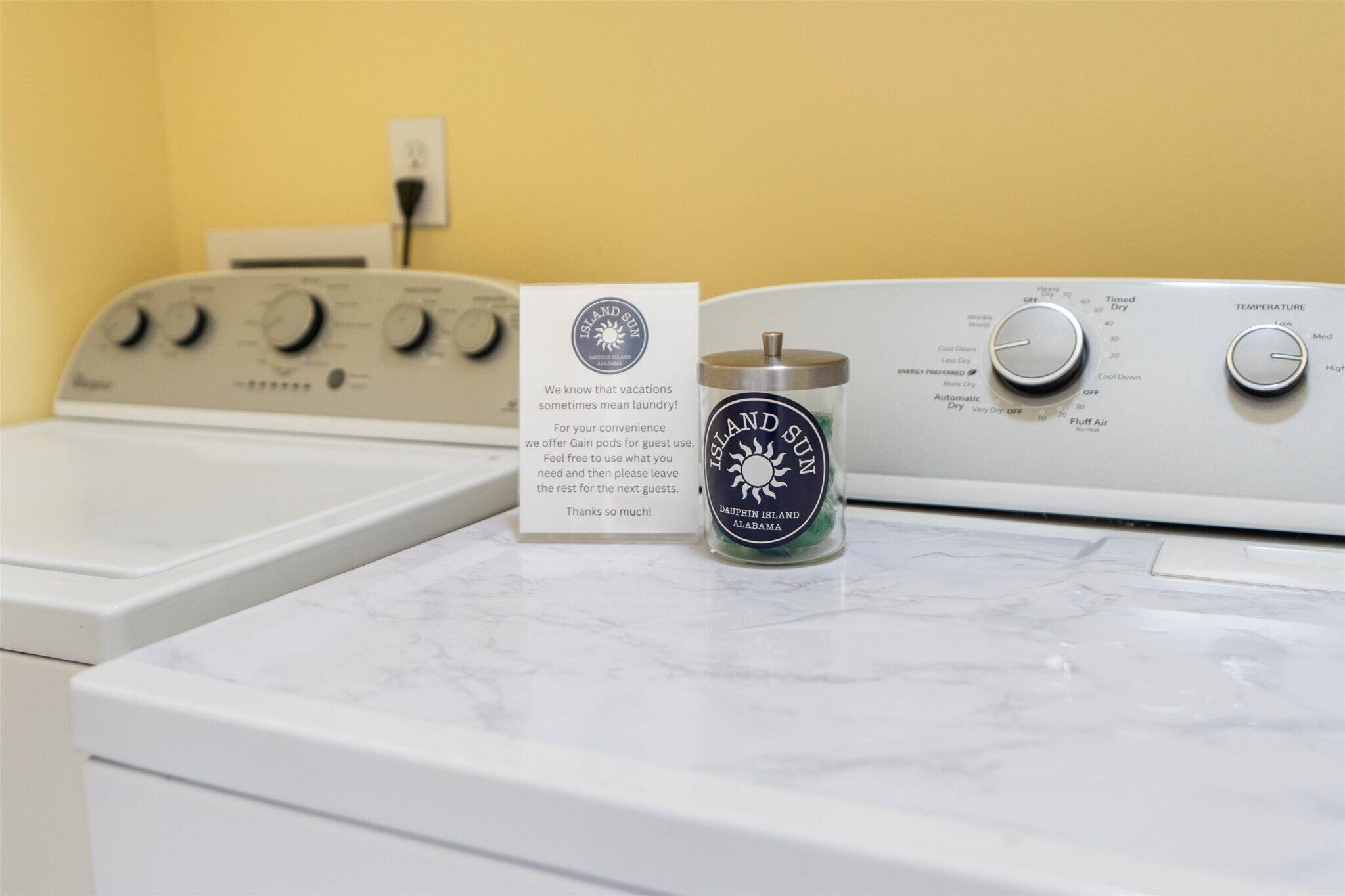 A front-loading washing machine with a control panel, a hand soap dispenser, and a card offering Gain pods for guest use.