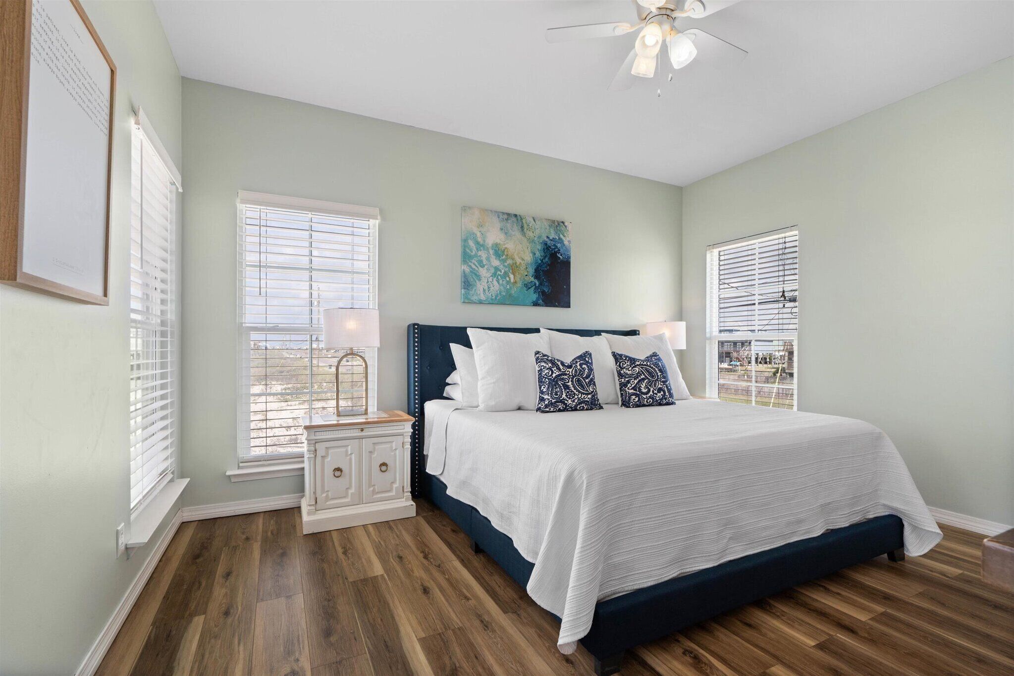 A bedroom with a large bed, white bedding, and a ceiling fan. There are two windows with blinds, a nightstand with a lamp, and a painting on the wall.