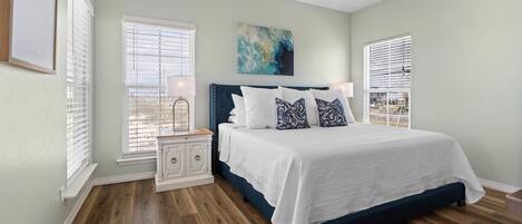 A bedroom with a large bed, white bedding, and a ceiling fan. There are two windows with blinds, a nightstand with a lamp, and a painting on the wall.