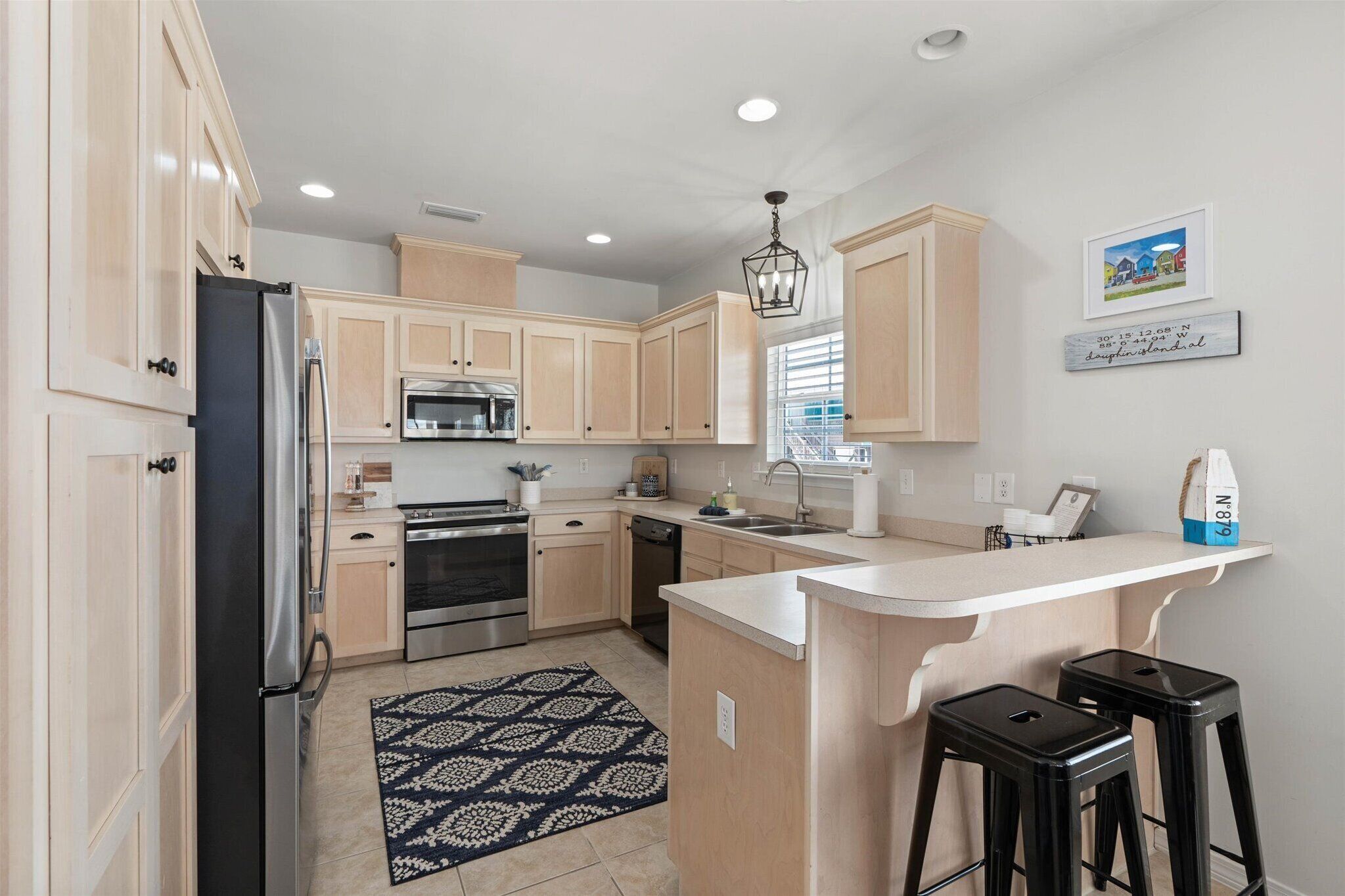 A modern kitchen with light wood cabinets, stainless steel appliances, a central island with stools, and a decorative rug.