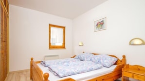 Une chambre individuelle avec un lit en bois, une fenêtre ornée d’un rideau blanc, une lampe fixée au mur et une petite table de chevet.