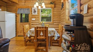 A wooden cabin interior with a dining table, chairs, a refrigerator, a coffee maker, and a window with a view of trees. - Little Joe Log Cabin, UTK/Neyland, Smokies, 16acres, FP, HotTub, Fishing, Pets (Knoxville)