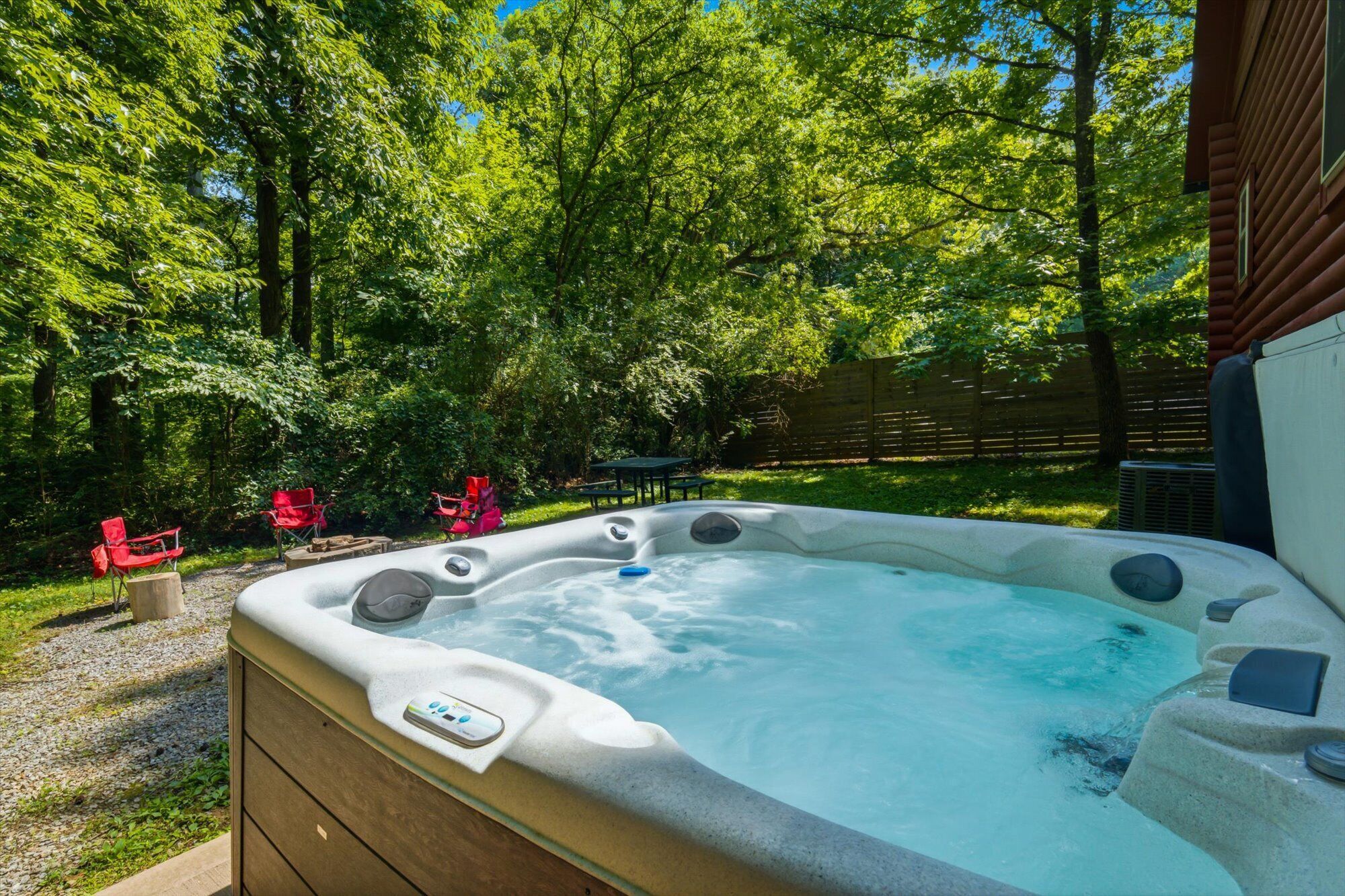 A hot tub in a backyard with a wooden deck, surrounded by trees and red chairs.
