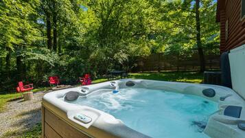 A hot tub in a backyard with a wooden deck, surrounded by trees and red chairs.