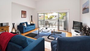 Una sala de estar moderna con un sofá azul, una mesa de centro y un televisor. Hay una puerta de cristal que da a un balcón con vistas a casas.