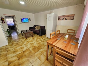 Una sala de estar con un sofá, una mesa de centro, una mesa de comedor con sillas, un refrigerador, un televisor y un cuadro en la pared.