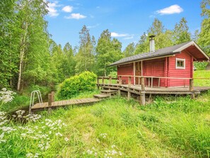 A red cabin with a wooden deck and a small bridge leading to it, surrounded by lush greenery and trees.