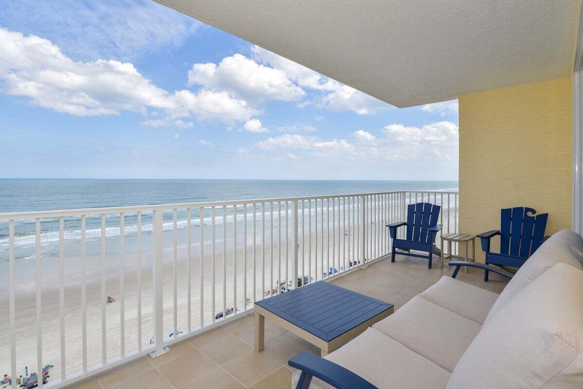 Een balkon met uitzicht op het strand en de oceaan. Er staan twee blauwe stoelen en een klein tafeltje.