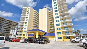 A multi-story hotel with a yellow facade, a blue-roofed entrance, and a parking lot with various vehicles. - High Tide Haven at the Aruba (Daytona Beach)