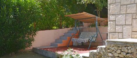 Un patio con sillón mecedora y zona de estar cubierta, rodeado de vegetación y un muro de piedra.