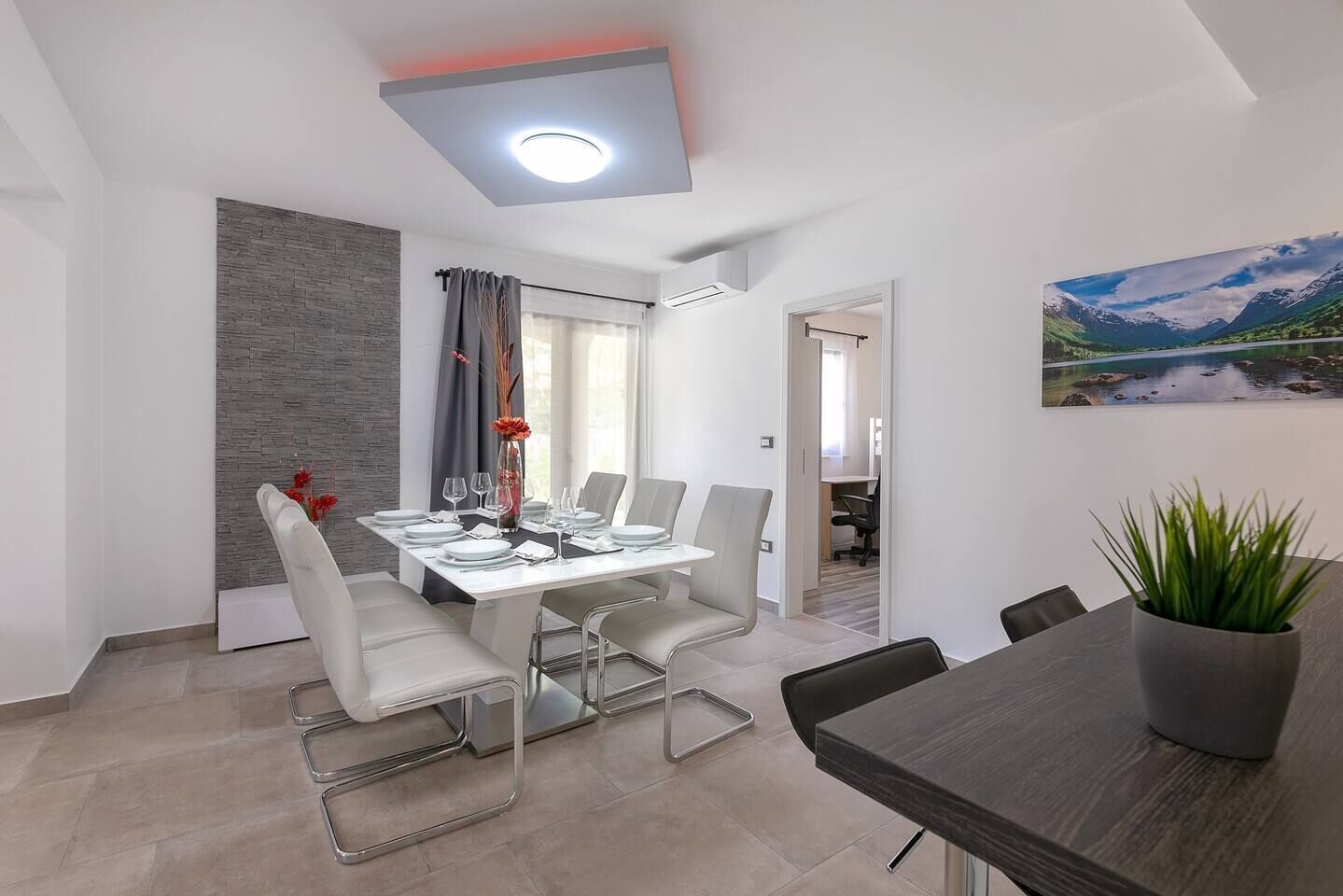 Et moderne spiseområde med et rektangulært bord dækket til fire personer, omgivet af hvide stole. En potteplante er placeret til højre, og et landskabsmaleri hænger på væggen.