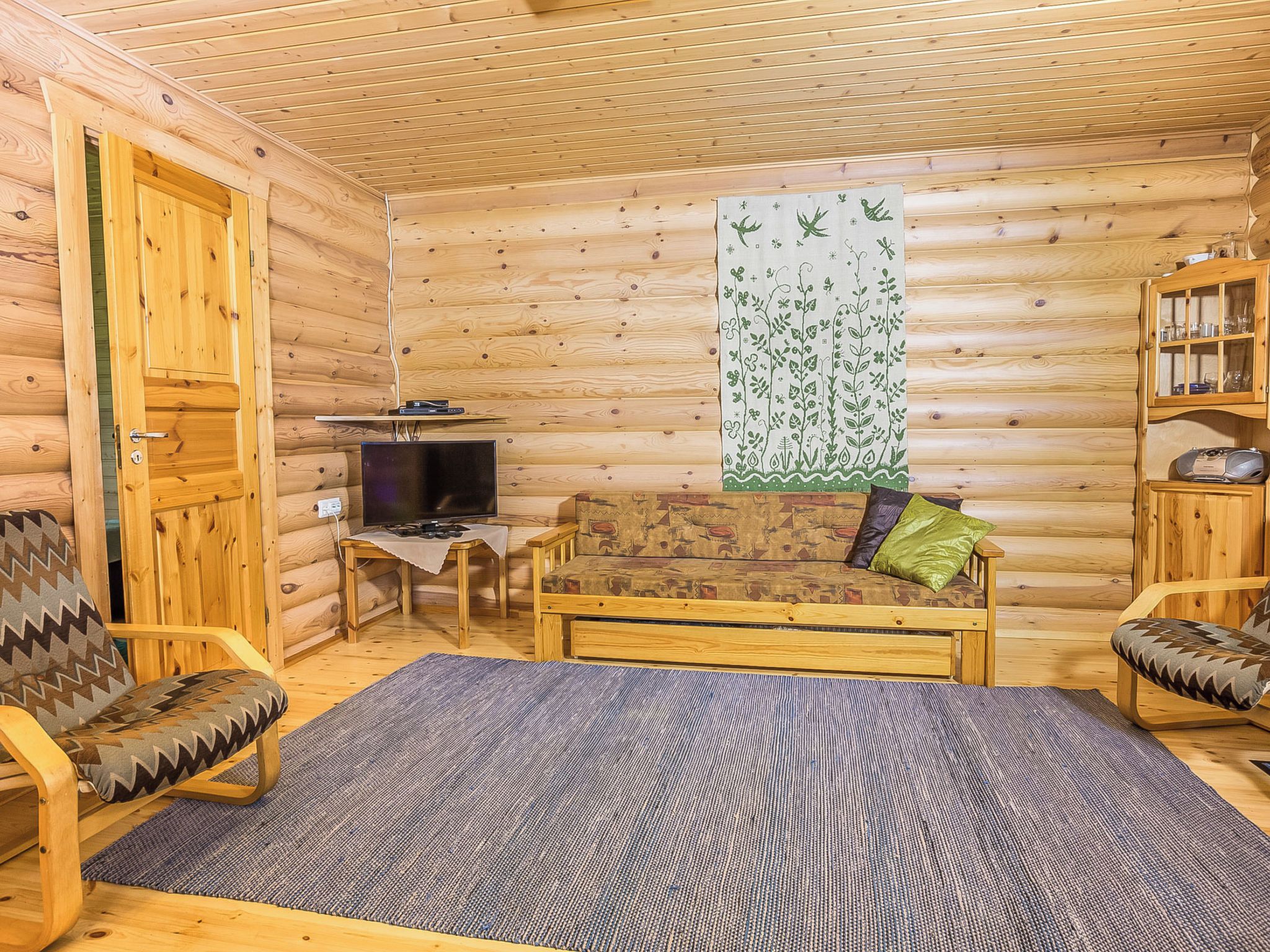 Ein Holzblockhaus-Interieur mit einer Couch, zwei Sesseln, einem kleinen Tisch, einem Fernseher und einem Bücherregal.