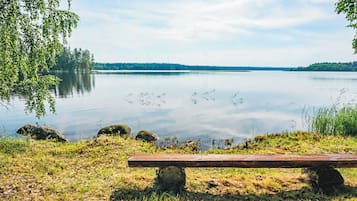 Een rustige oever met een houten bankje en rotsen op de voorgrond, een heldere lucht en een bos in de verte.