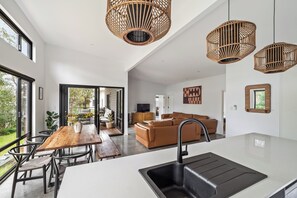 Una cocina moderna con una isla central, un comedor con una mesa y sillas de madera, una sala de estar con un sofá marrón, y un amplio ventanal con vistas al exterior.