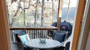 A covered outdoor seating area with wicker chairs and a round table, a welcome mat, and a view of trees and a building in the background.