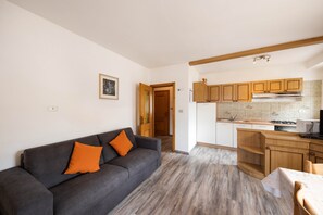 A living room with a sofa and a table, a kitchen with wooden cabinets and a stove, and a hallway leading to another room. - Appartamento Primavera 1 with Mountain View, Wi-Fi, Balcony & Garden (Moena)