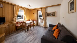 A room with a wooden floor, a sofa, a dining table, and a television. - Appartamento Primavera 1 with Mountain View, Wi-Fi, Balcony & Garden (Moena)
