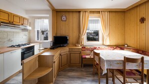 A compact kitchen with wooden cabinets, a gas stove, a built-in oven, a television, and a dining area with a bench and table. - Appartamento Primavera 1 with Mountain View, Wi-Fi, Balcony & Garden (Moena)