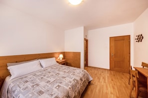 Un dormitorio con cabecera de madera, cama con ropa de cama estampada, mesita de noche con lámpara, puerta de madera y un pequeño escritorio de madera con silla.