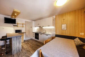 A compact kitchen with a microwave, stove, and sink, a small bar area with stools, a dining table with chairs, and a wooden wall with a cross.