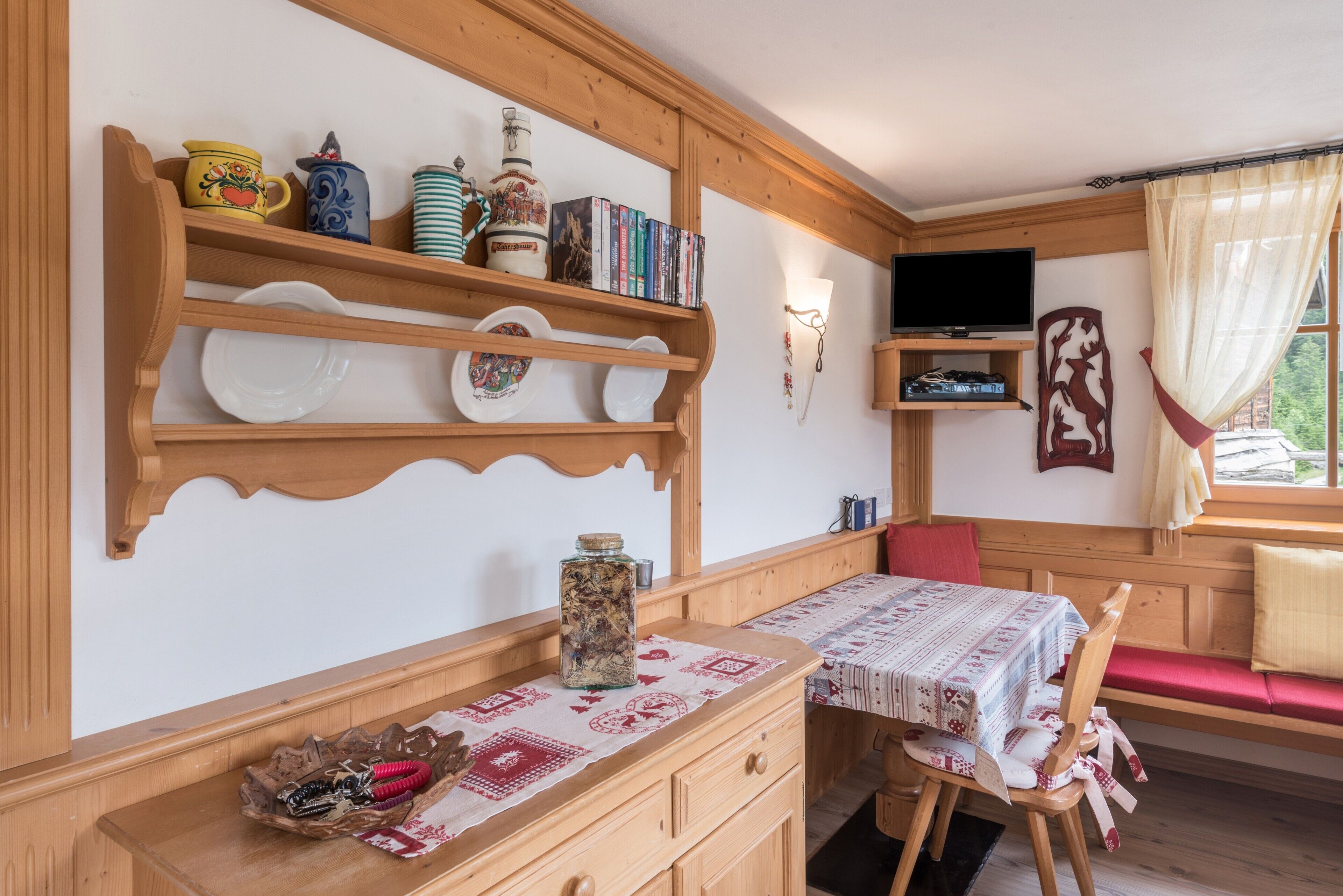 Une pièce chaleureuse avec des étagères en bois, une table recouverte d’une nappe à motifs, un téléviseur et une fenêtre avec des rideaux.