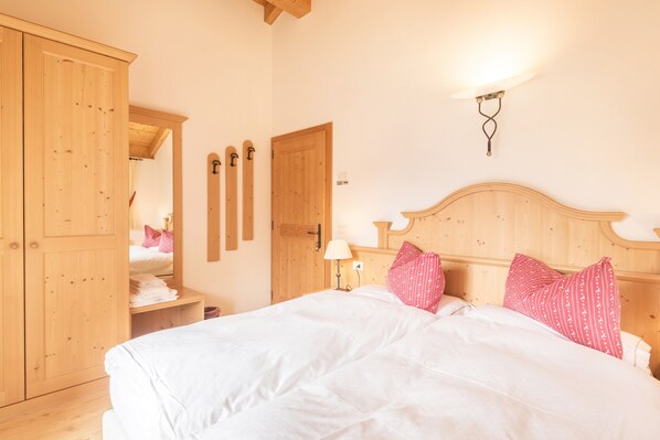 A bedroom with a wooden wardrobe, a bed with white bedding and pink pillows, a nightstand with a lamp, and a wall-mounted light fixture.