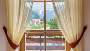 A window with curtains, a view of a mountain, and a building with a red roof. - Vacation Chalet Crepes De Sela with Mountain View, Wi-Fi, Balcony & Garden (Canazei)