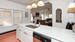 A modern kitchen with white cabinetry, a built-in dishwasher, and a white countertop. There is a window with blinds, a dining area with a table and chairs, and a ceiling fan. - Close to Downtown, LECOM Field, AMI Beaches and more (Bradenton)