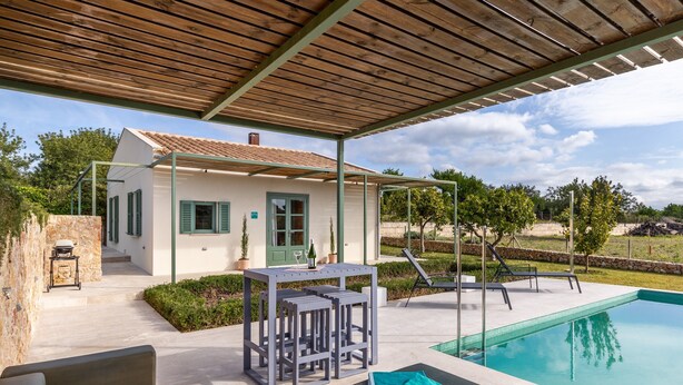 Un espacio al aire libre techado con piscina, una mesa con sillas y un pequeño edificio con puerta y ventanas.