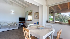 Un área de comedor con una mesa, sillas, un plato de comida, un televisor y un sofá en una habitación luminosa con una ventana.