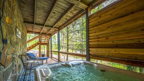 Un jacuzzi en una cabaña de madera con pared de piedra, una silla y vistas a árboles.