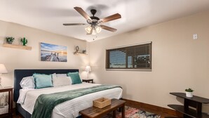 Una camera da letto con un letto, una ventola a soffitto, una finestra con tende, un comodino, una lampada, una decorazione con un cactus e un quadro appeso al muro.