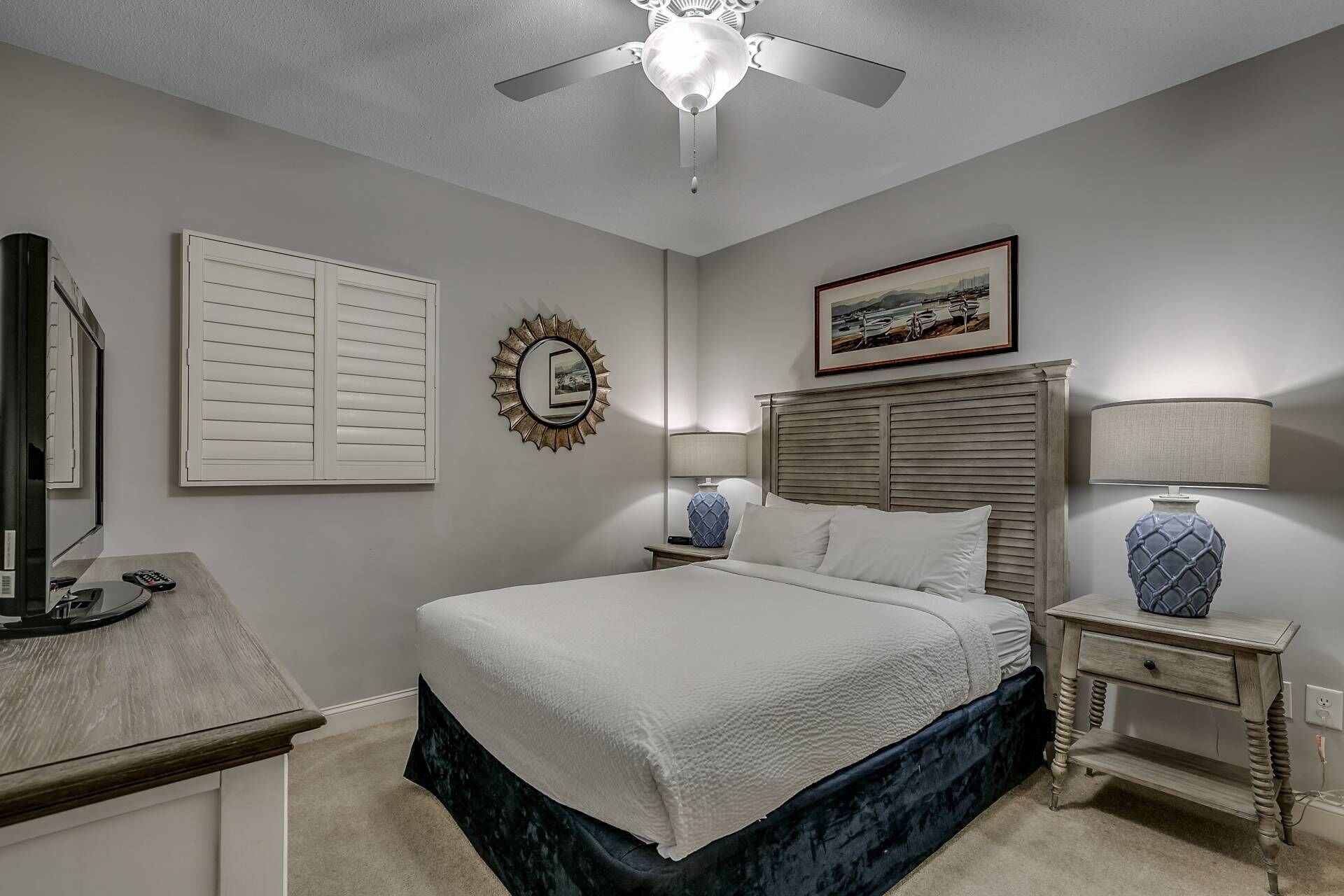 A bedroom with a bed, a ceiling fan, a nightstand with a lamp, a framed picture on the wall, and a window with blinds.