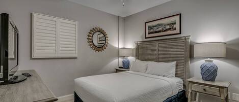 A bedroom with a bed, a ceiling fan, a nightstand with a lamp, a framed picture on the wall, and a window with blinds.