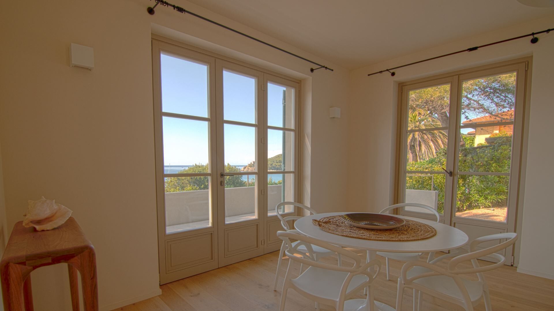 Une pièce avec une table ronde et quatre chaises, de grandes fenêtres donnant sur la mer et la verdure, et un tabouret en bois.
