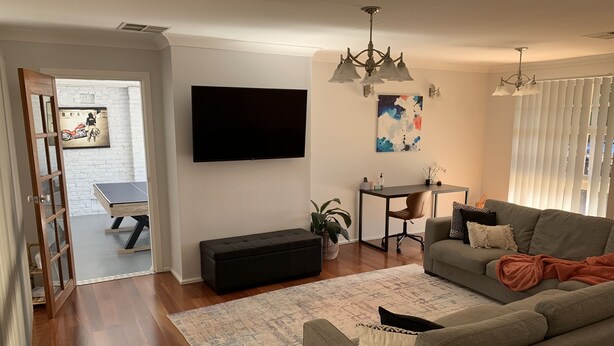 Un salon avec un canapé gris, un pouf noir, une table basse, un téléviseur, un bureau avec une chaise et un tableau accroché au mur.