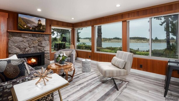 A cozy living room with a stone fireplace, comfortable seating, and large windows overlooking a lake. - *New Listing* Beautiful Riverfront Cottage, Newly Remodeled with Amazing Views, BBQ, Close to Town. (Florence)