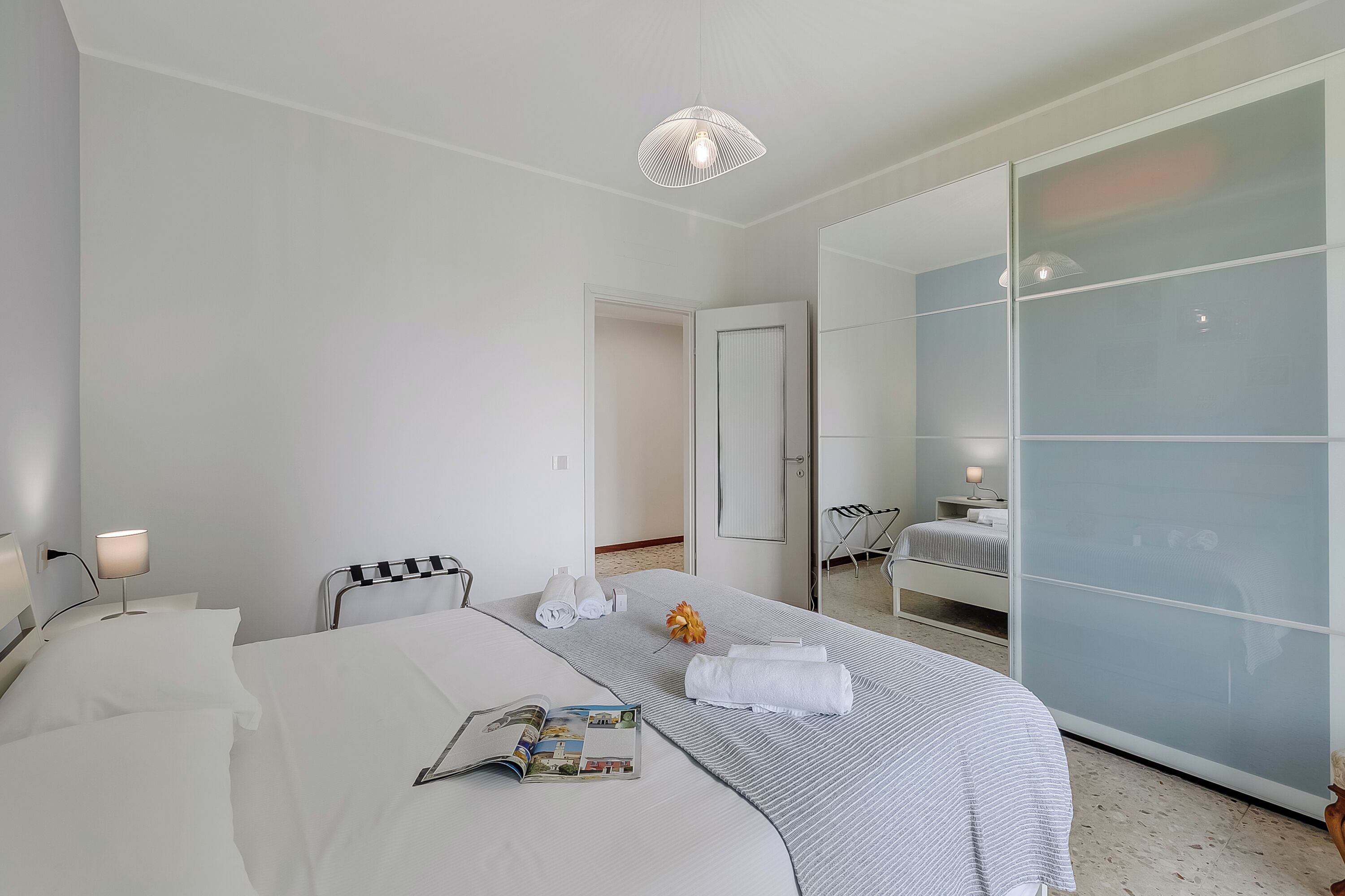 Habitación de hotel moderna con una cama grande, una mesita de noche con lámpara, un espejo y un divisorio de vidrio corredero que da a un baño.