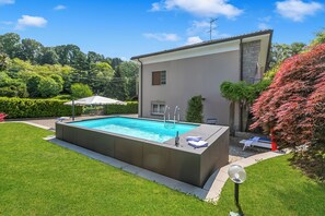 A rectangular swimming pool with a built-in deck, surrounded by a well-maintained lawn and a house with a stone facade. - Villa Giulietta Private Pool (Gemonio)