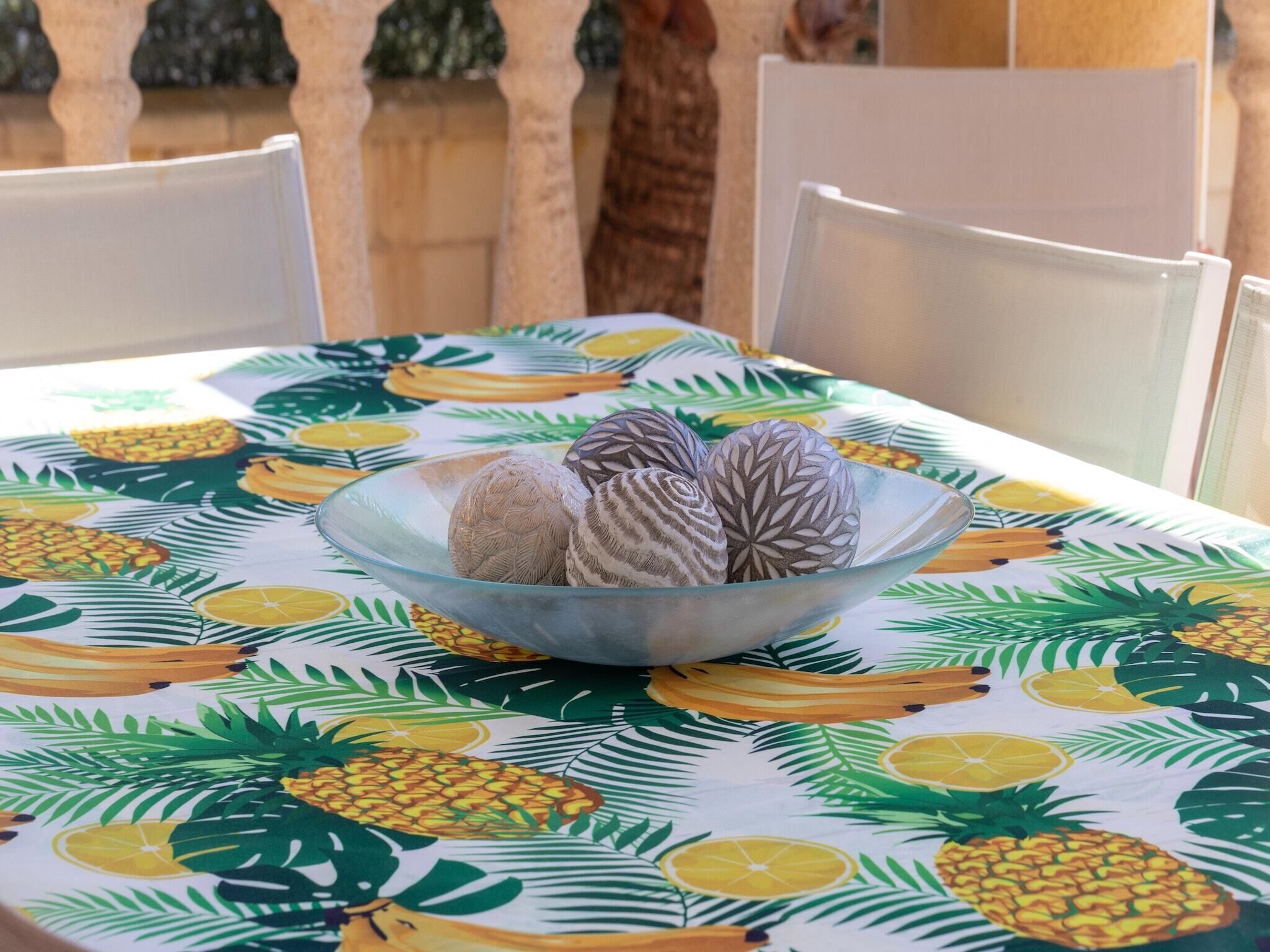 A table with a fruit-themed tablecloth, a glass bowl with decorative items, and white chairs.