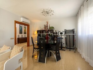 Een eethoek met een glazen tafel, stoelen, een rode vaas met gedroogde bloemen, een plafondlamp en een loopband.