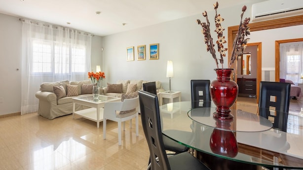 Un salon moderne avec une table à manger en verre, des canapés beige, un vase rouge et un coin repas avec des chaises.