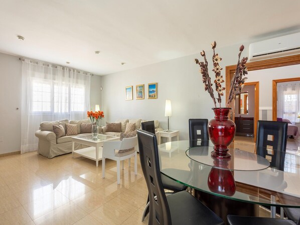 A modern living room with a glass dining table, beige sofas, a red vase, and a dining area with chairs.
