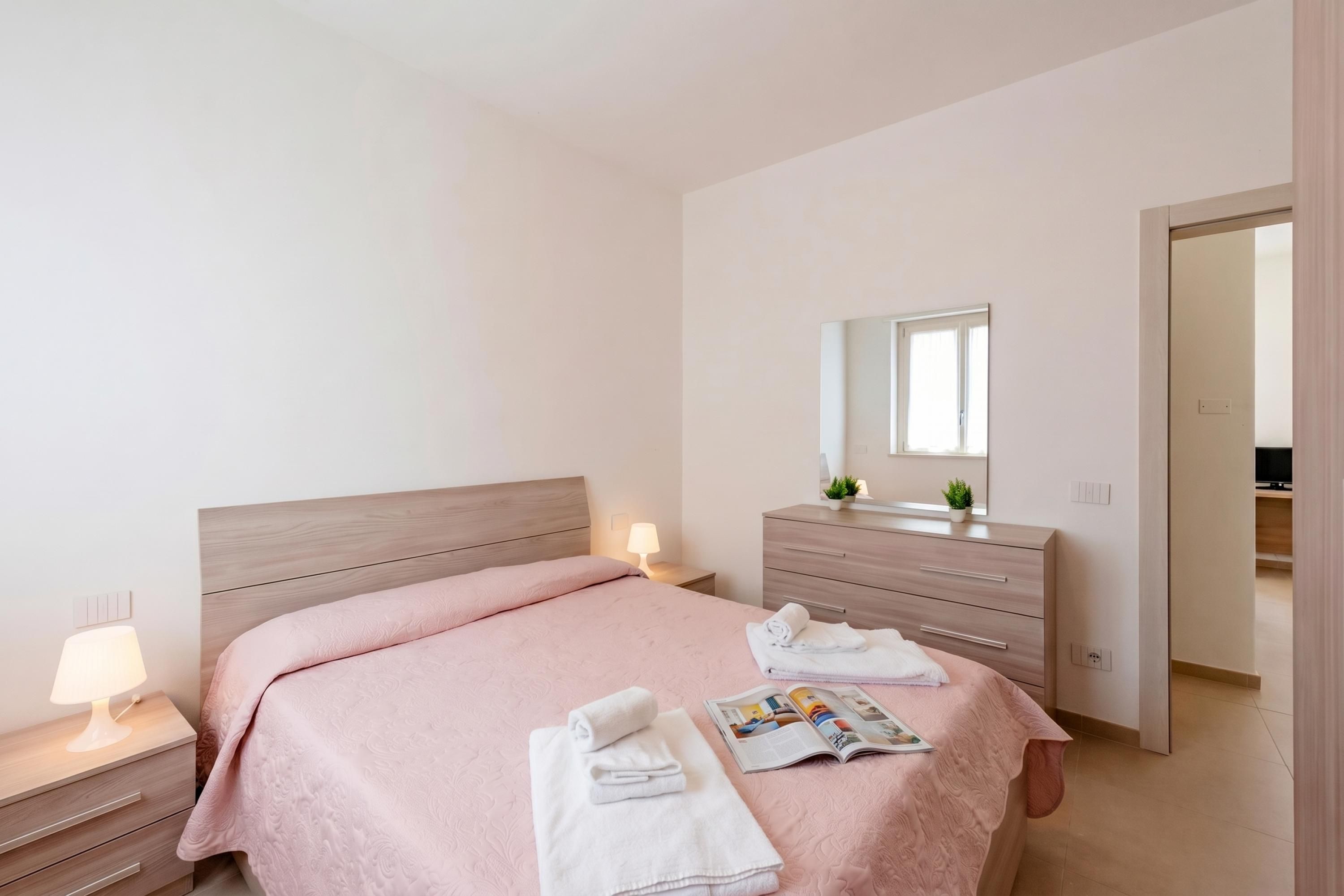 A modern bedroom with a wooden headboard, a bedside table with a lamp, a dresser with a mirror, and a magazine on the bed.