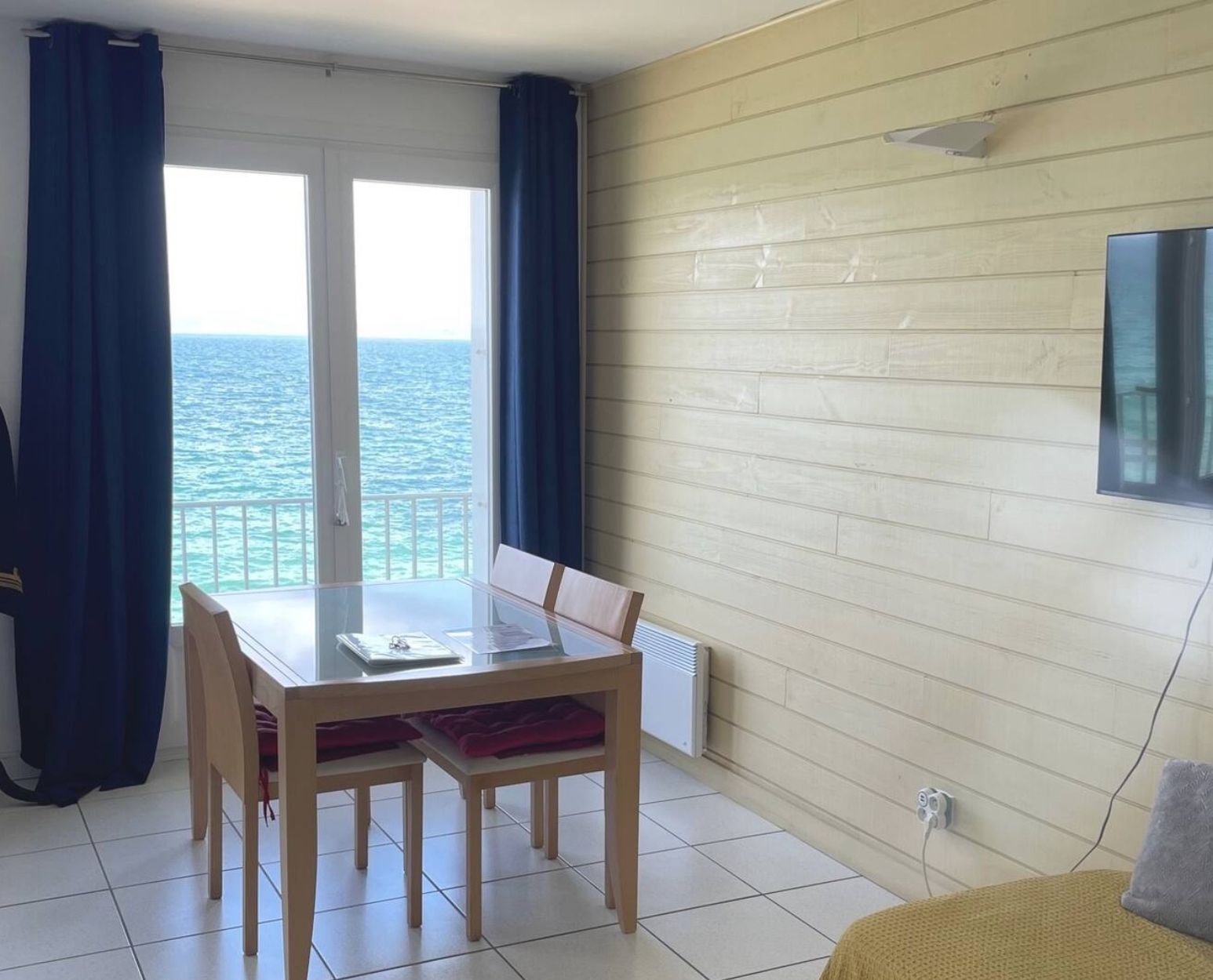 Een kamer met een tafel en stoelen, uitzicht op zee door een glazen deur, een wandlamp en een stopcontact.