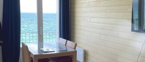 A room with a table and chairs, a view of the sea through a glass door, a wall-mounted light, and a power outlet.
