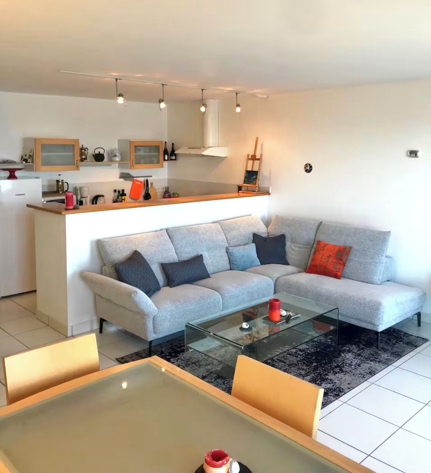 Ein modernes Wohnzimmer mit einem grauen Sofa, einem Glastisch und einer Küchenzeile im Hintergrund.