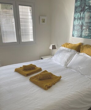 Uma cama bem arrumada com roupa de cama branca e amarela, um abajur e um quadro na parede.