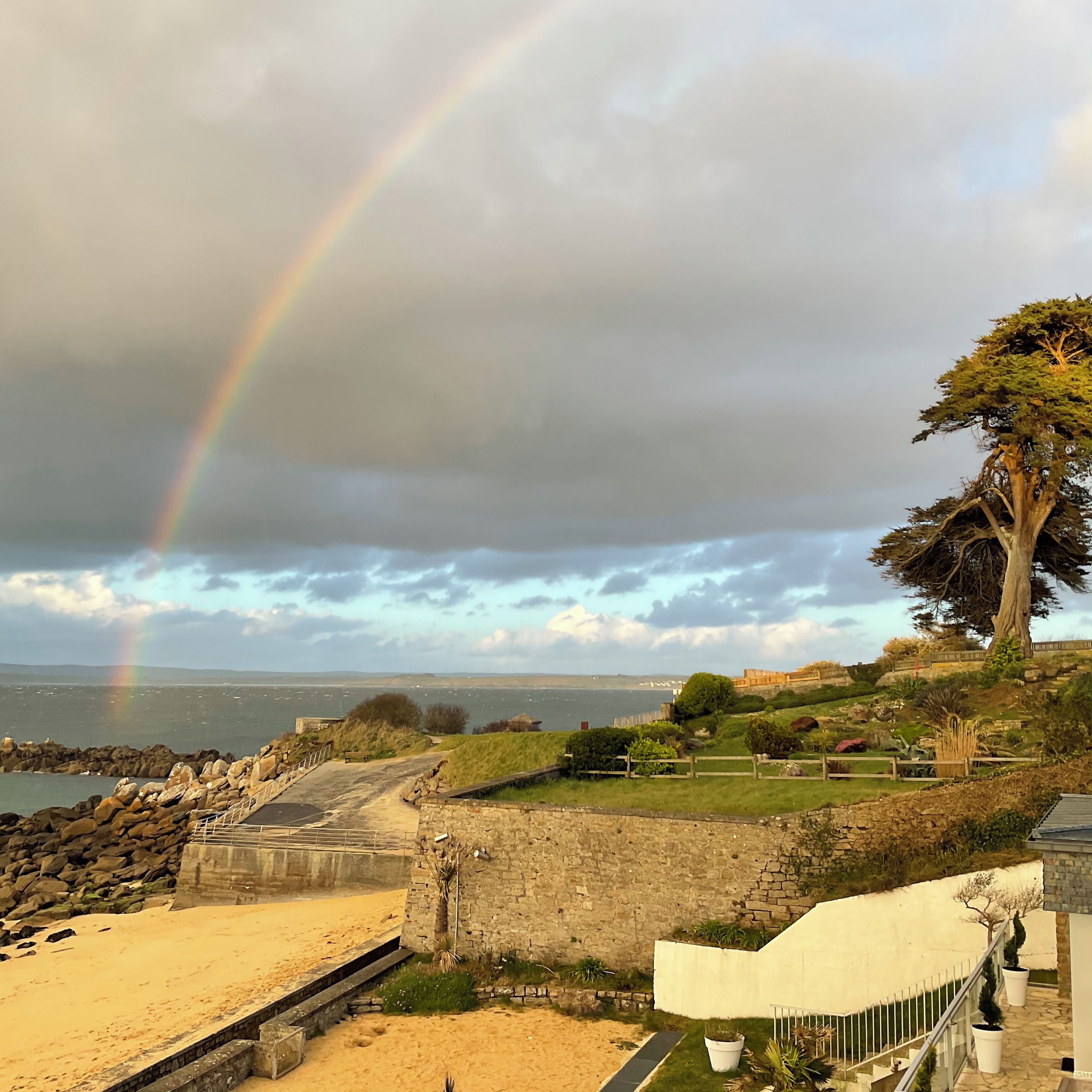 A coastal landscape with a rainbow, a sandy beach, and a stone wall.