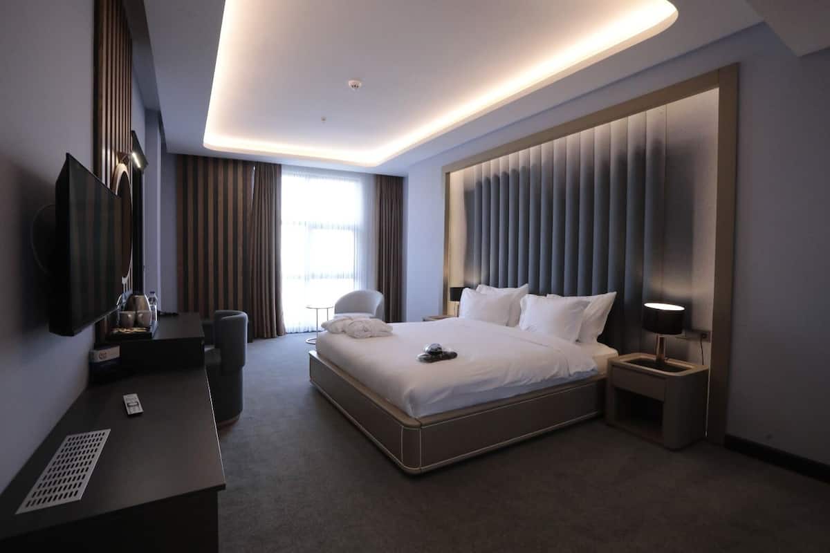 A modern hotel room with a large bed, a desk, a TV, and a bedside table with a lamp.